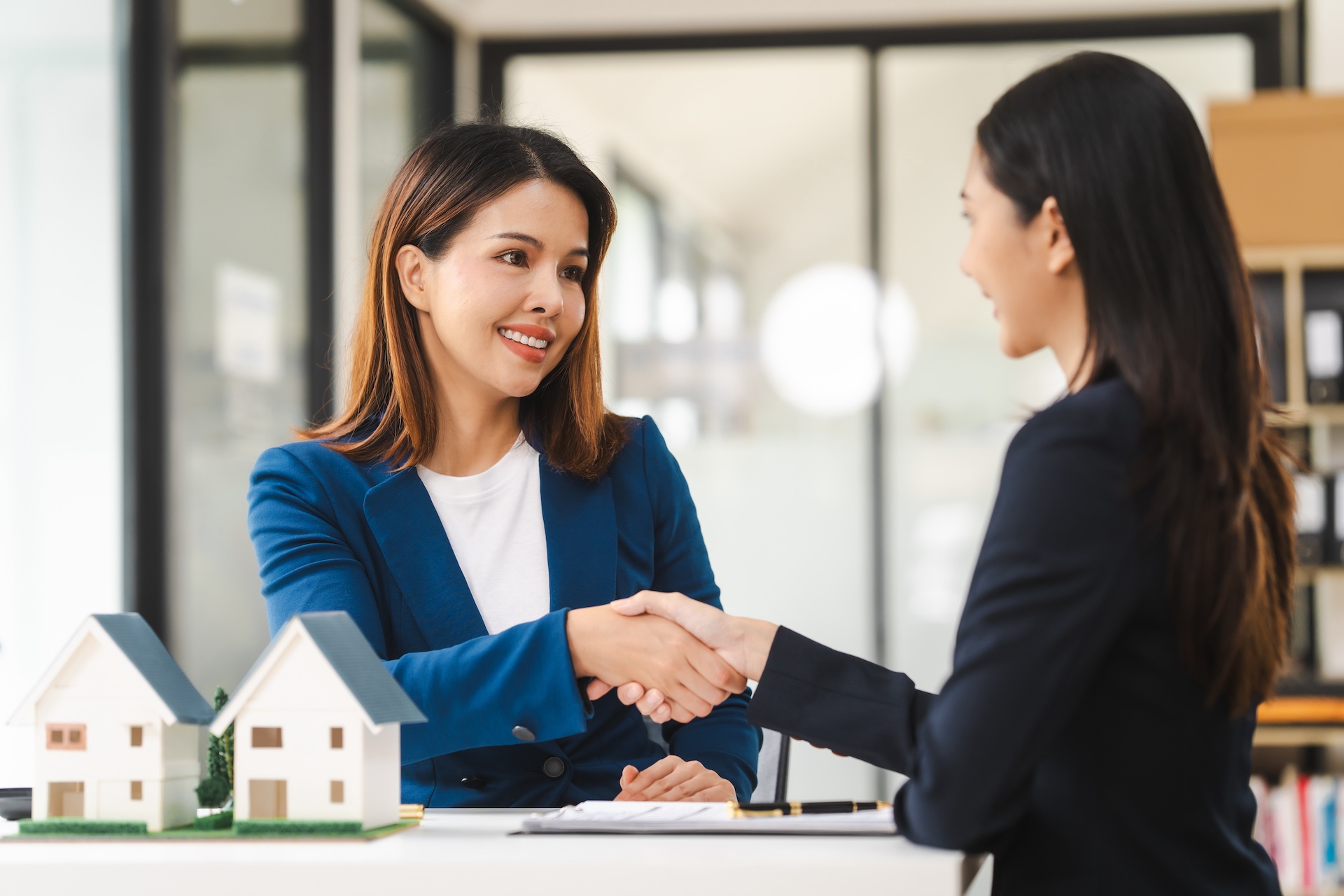 Two female real estate agents in formal suits discuss a contract with house models on the table, ready to sign a deal. Represents one agent recruiting another to her real estate brokerage network for revenue streams opportunities.